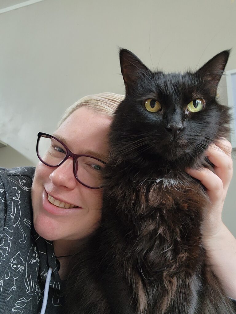 Tory (a white female with glasses and short blonde hair) smiles while resting her head against her fluffy black cat Mandy.